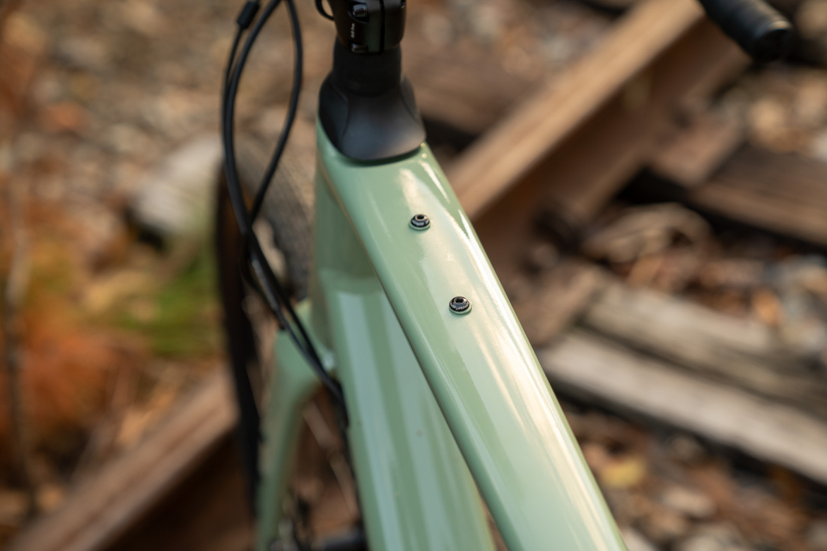 Close-up of a pale green bicycle frame, highlighting two mounting bolts on the top tube. The background features blurred railroad ties and natural foliage, suggesting an outdoor setting.