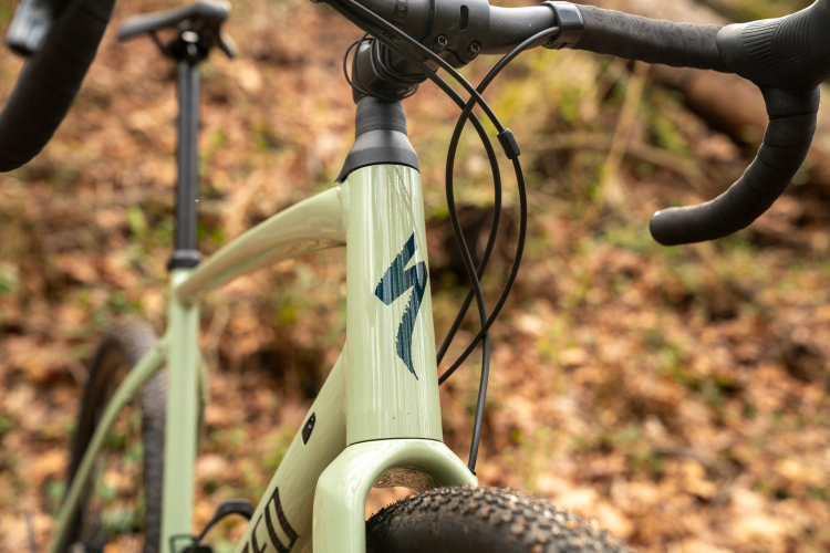 Close-up of a light green bicycle frame with a prominent logo, showcasing the handlebars and cables against a blurred natural background of brown leaves and greenery.
