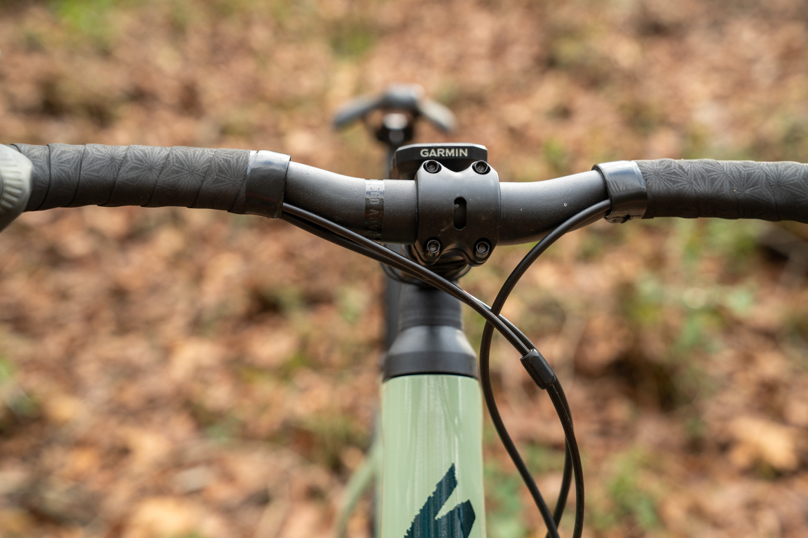 Close-up view of a bicycle handlebar and stem, featuring textured black grips and a Garmin mount, with a blurred natural background of leaves.