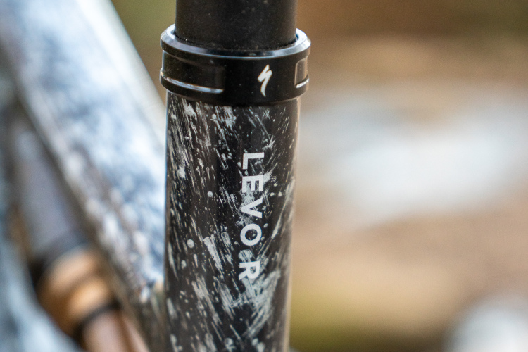 Close-up image of a bike's seat post with visible dirt and scratches, featuring the brand name "LEVOR" and a small logo. The background is softly blurred, suggesting an outdoor setting.