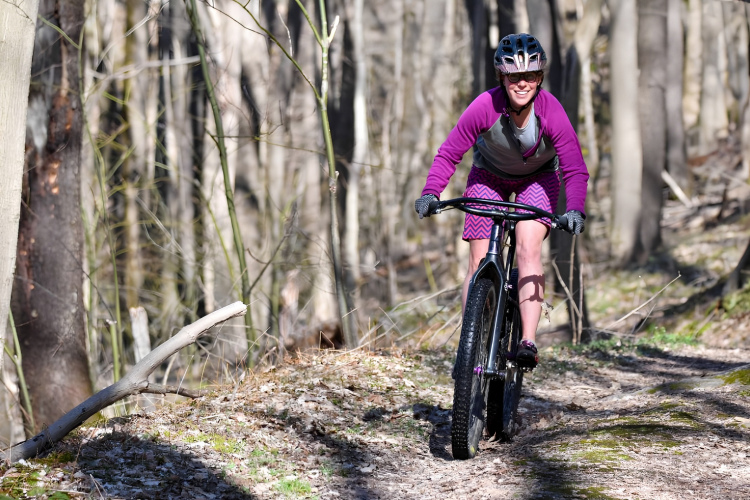 A smiling individual wearing a purple long-sleeve top and patterned shorts rides a mountain bike along a dirt trail surrounded by trees in a forest. The sun filters through the branches, illuminating the scene as they navigate the pathway.