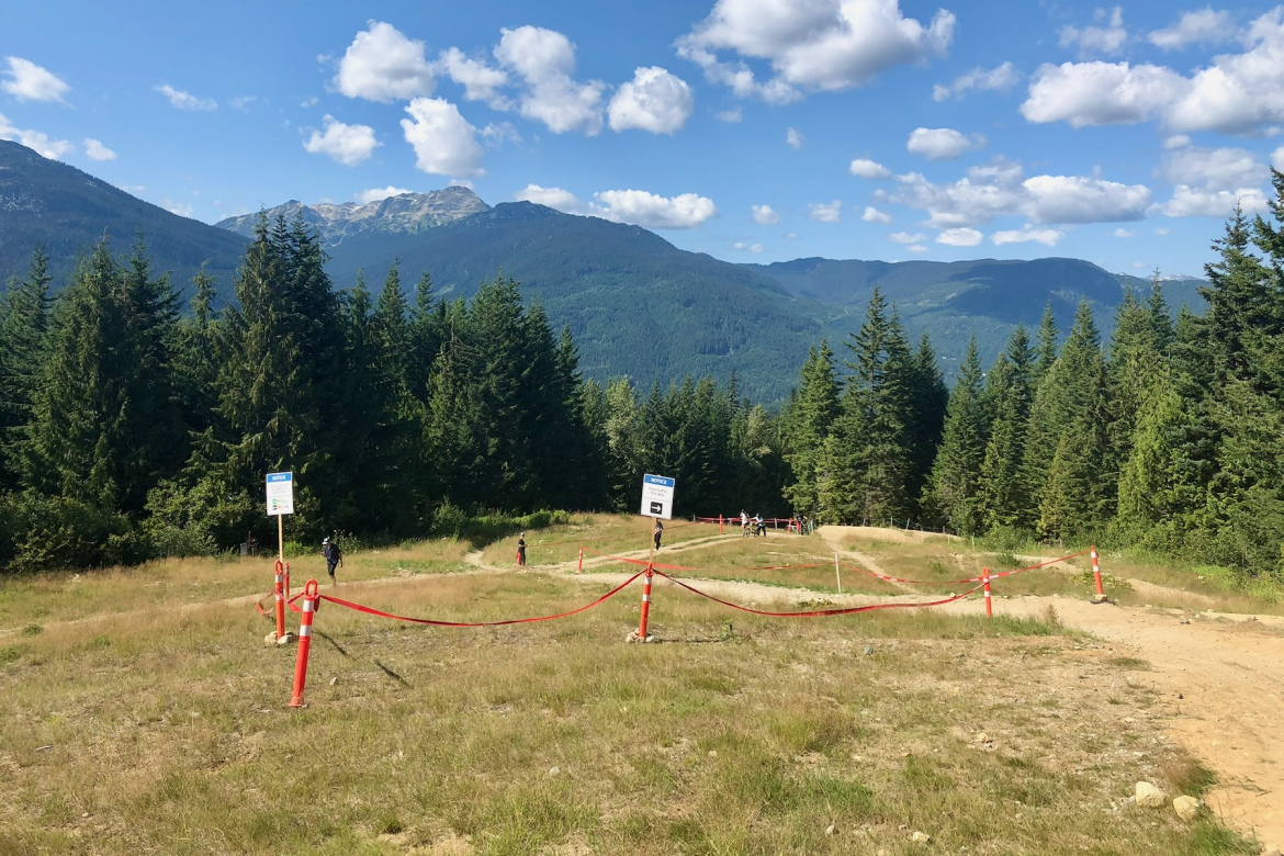 A mountain biking trail at Whistler Bike Park surrounded by lush green trees and distant mountains, with a clear blue sky dotted with fluffy clouds. Red safety markers and a sign are visible along the trail, and a few people can be seen walking or biking on the path.