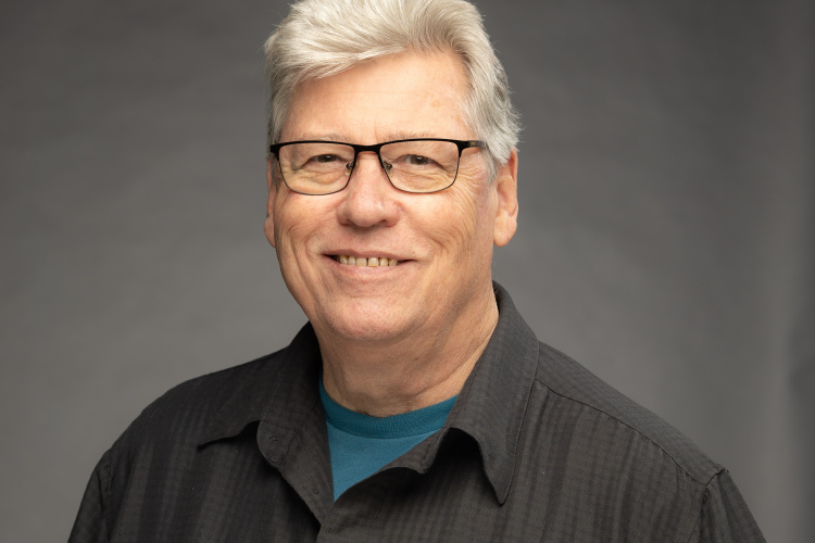 A smiling older man with grey hair and glasses, wearing a black collared shirt over a blue shirt, poses against a neutral grey background.