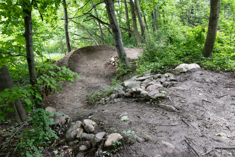 A dirt biking trail curving through a lush green wooded area, with trees lining the path and rocks forming a border along the edge. The trail is well-maintained, showing signs of recent use, and the surrounding foliage includes various plants and underbrush.