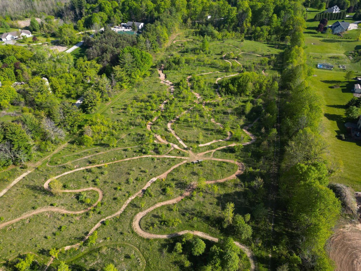Aerial view of a winding Rocky Knoll trail system surrounded by lush greenery, with residential homes visible in the background. The landscape features various paths that meander through a densely wooded area, highlighting the natural terrain and diverse vegetation.