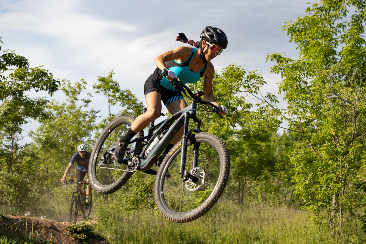 A mountain biker performs a jump on a dirt trail, surrounded by greenery. The rider is wearing a blue tank top, black shorts, and a helmet, showcasing an athletic posture as they soar through the air. Another cyclist can be seen in the background, riding along the trail. Dust rises from the ground beneath them, capturing the dynamic energy of the activity.