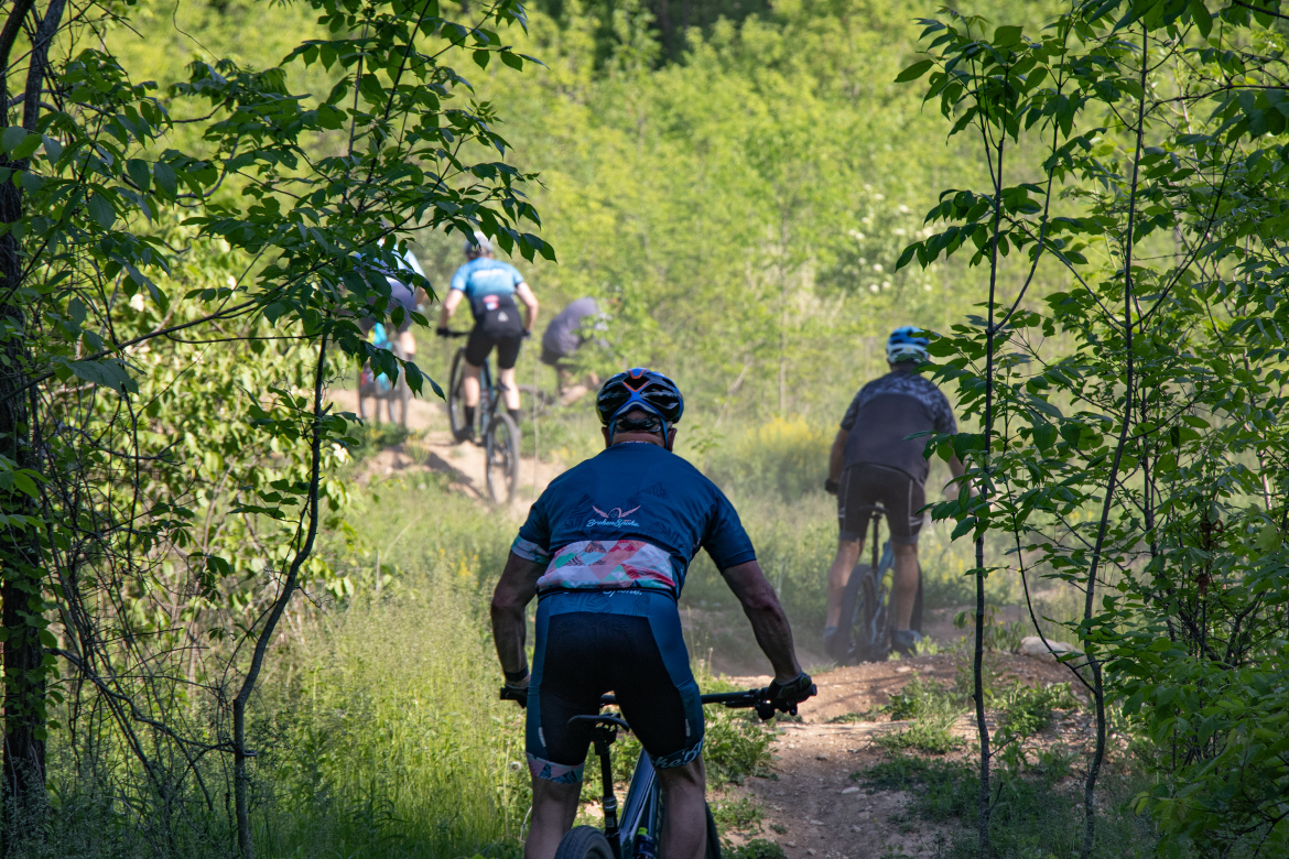A group of mountain bikers riding on a dusty trail through a lush green forest at Rocky Knoll Trail System. In the foreground, a cyclist in a blue jersey and black shorts is seen from behind, with several other bikers visible in the background navigating the path. The scene captures the vibrant greenery and the sense of adventure in nature.