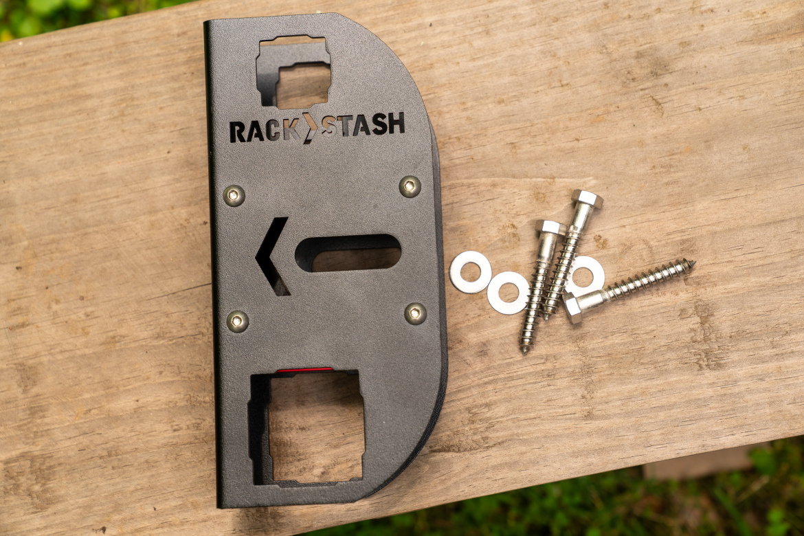 A metal tool marked with "RACK & STASH" on a wooden surface, accompanied by screws and washers. The tool features cutouts and mounting points, suggesting it is used for assembly or installation purposes.