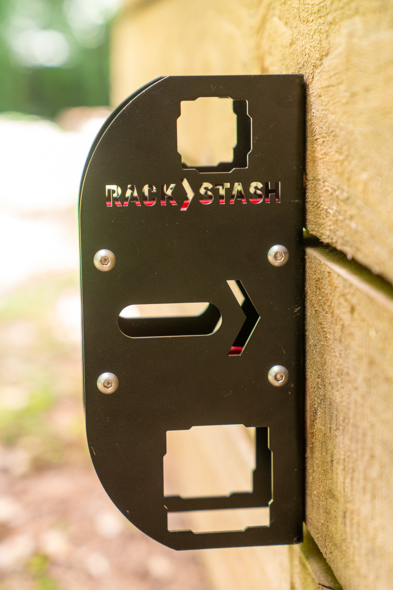 Metal bracket mounted on a wooden surface, featuring the words "RACK STASH" cut into the material. The bracket has an arrow design and multiple cut-out shapes.