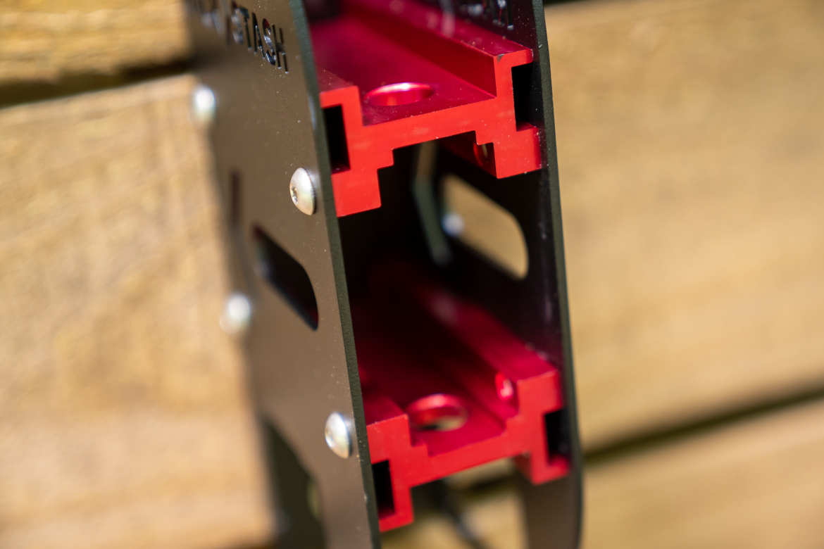 Close-up of a black metal tool or fixture featuring red components inside, set against a wooden background. The image shows intricate detailing, including openings and screws, highlighting its mechanical design.