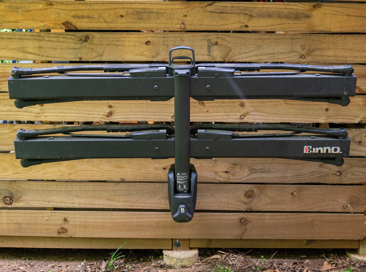A black bike rack mounted on a wooden fence, featuring two horizontal arms for holding bicycles. The rack has a handle at the top and is installed securely into the ground. The background shows natural wood grain and a few green plants.