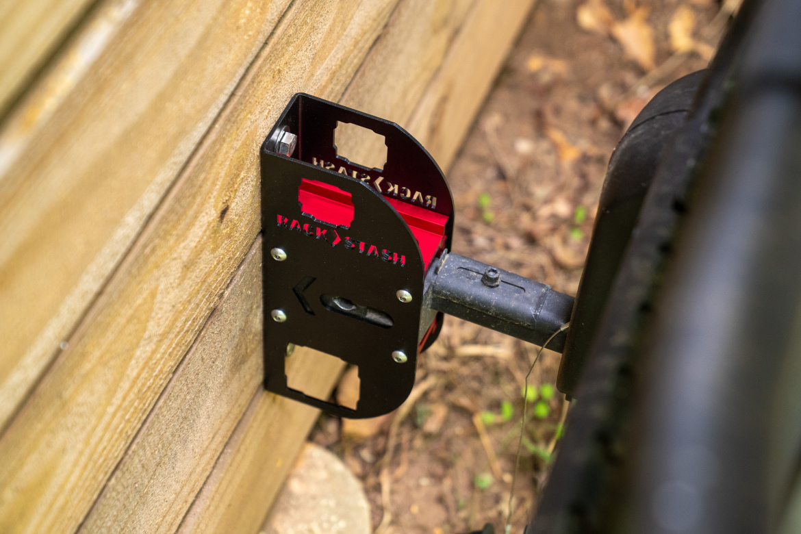 A close-up view of a black and red rack attachment mounted on a wooden surface, designed for outdoor use. The attachment features cutouts for securing equipment and is connected to a cylindrical component, with earth and foliage visible in the background.