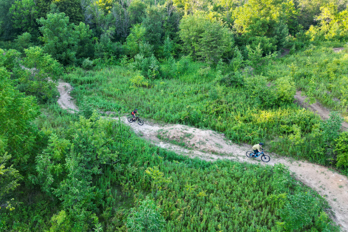 Aerial view of two mountain bikers riding along a winding trail at Rocky Knoll Trail System through lush greenery in a wooded area. The surrounding vegetation includes dense trees and tall grass, suggesting a vibrant outdoor environment.