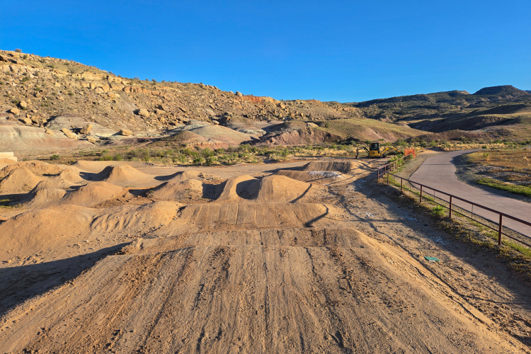 A dirt bike park featuring a series of dirt jumps and mounds, set against a backdrop of rocky hills and a clear blue sky. A paved path is visible on the right side, alongside a fenced area, and a piece of construction equipment can be seen in the distance. The landscape is dry and sandy, characteristic of an outdoor recreational area.