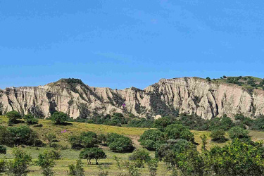 Landscape featuring a rocky outcrop with steep, layered cliffs under a clear blue sky. The foreground consists of grassy meadows dotted with small shrubs and wildflowers. The scene conveys a serene natural setting. Vashlovani National Park Adventure Loop mountain bike trail.