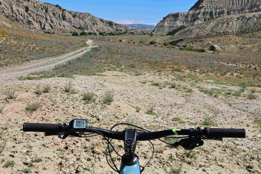 A view from the handlebars of a mountain bike, overlooking a rugged, arid landscape with hills and a clear blue sky. A dirt path winds through the terrain, bordered by sparse vegetation. Vashlovani National Park Adventure Loop mountain bike trail.