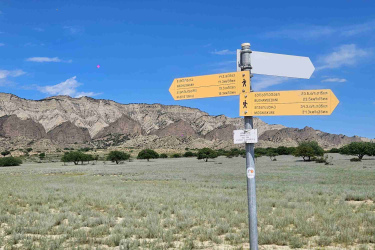 A signpost with multiple directional arrows, featuring yellow and white signs indicating various destinations and distances, set against a clear blue sky and rugged mountainous landscape in the background. The foreground shows a flat, grassy area with sparse vegetation. Vashlovani National Park Adventure Loop mountain bike trail.