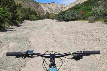 A view from the handlebars of a mountain bike on a sandy trail in a rocky canyon landscape, with blue skies and greenery on the sides. Vashlovani National Park Adventure Loop mountain bike trail.
