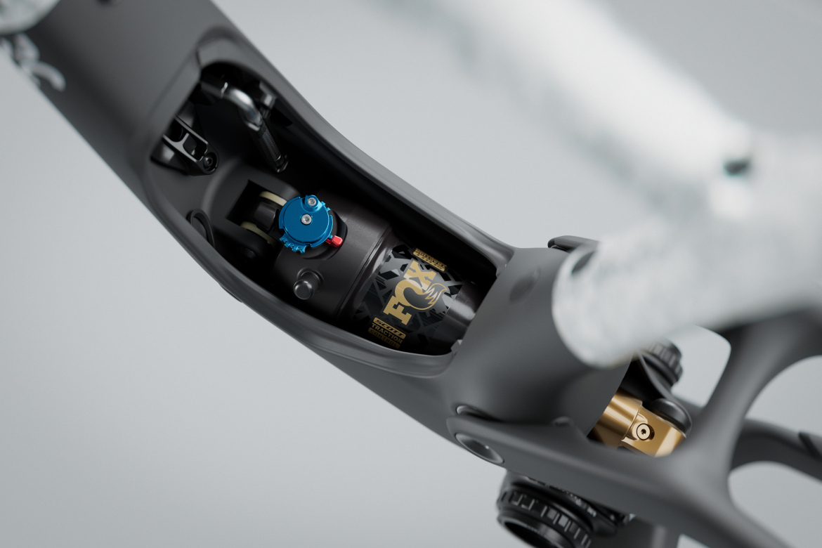 Close-up view of a bicycle frame showcasing the internal suspension system, featuring a FOX shock absorber with a blue adjustment knob and gold branding, set against a blurred gray background.