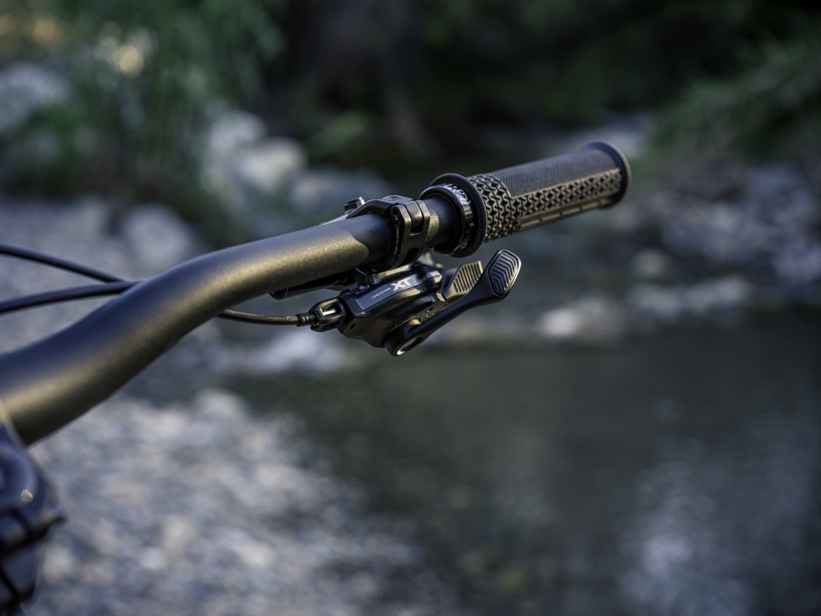 Close-up view of a bicycle handlebar featuring textured grips and a gear shifter, set against a blurred natural background of rocks and greenery.