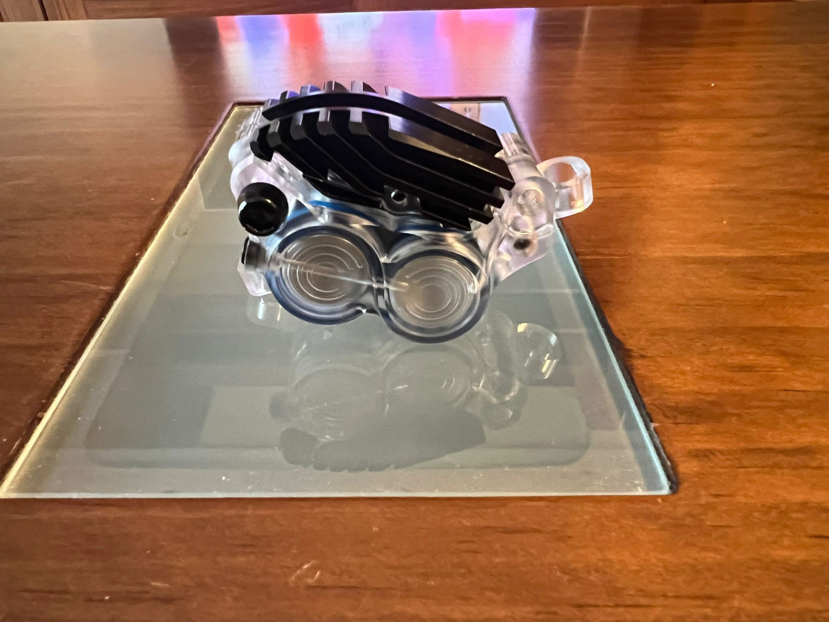 A transparent Hustok brake caliper with a black, textured top is positioned on a glass surface. The object features two circular elements at the base, and it reflects light, creating a slight shine. The background consists of a wooden surface, adding warmth to the overall composition.