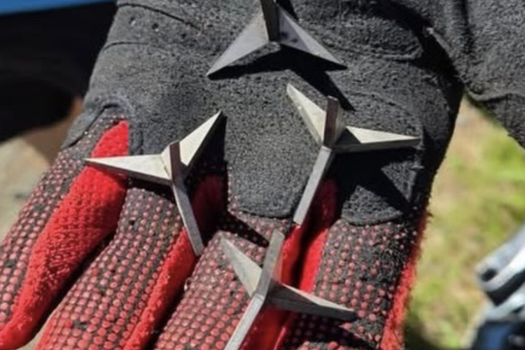 A close-up of a hand wearing a black glove with red detailing, holding four triangular metal objects. The background features blurred greenery and a hint of a vehicle.