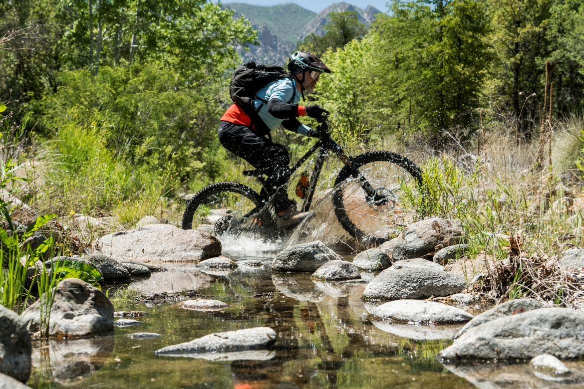 A mountain biker splashes through a shallow stream while navigating rocky terrain in a lush green environment. The cyclist is wearing a helmet and protective gear, with water droplets flying around as they pedal over the stones. Sunlight filters through the trees, highlighting the vibrant scenery.