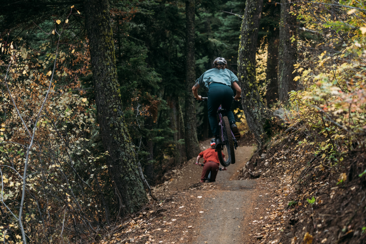 A cyclist in a green shirt and black pants rides a mountain bike along a dirt trail, performing a jump. A child in a red outfit is biking closely behind. The scene is surrounded by trees with autumn foliage, creating a vibrant and natural setting.