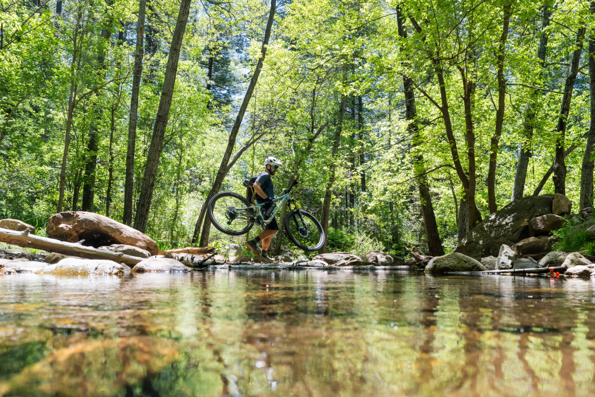 A person carrying a mountain bike while crossing a shallow stream in a lush, green forest. Sunlight filters through the trees, illuminating the vibrant foliage and reflecting off the water's surface. Highline Trail: Geronimo's Call mountain bike trail.