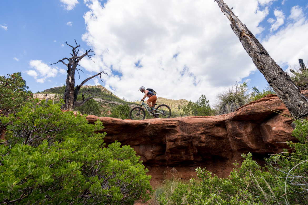 A mountain biker navigating a rocky ledge surrounded by greenery, with a mountainous landscape and cloudy blue sky in the background. Highline Trail: Inner Sanctum mountain bike trail.