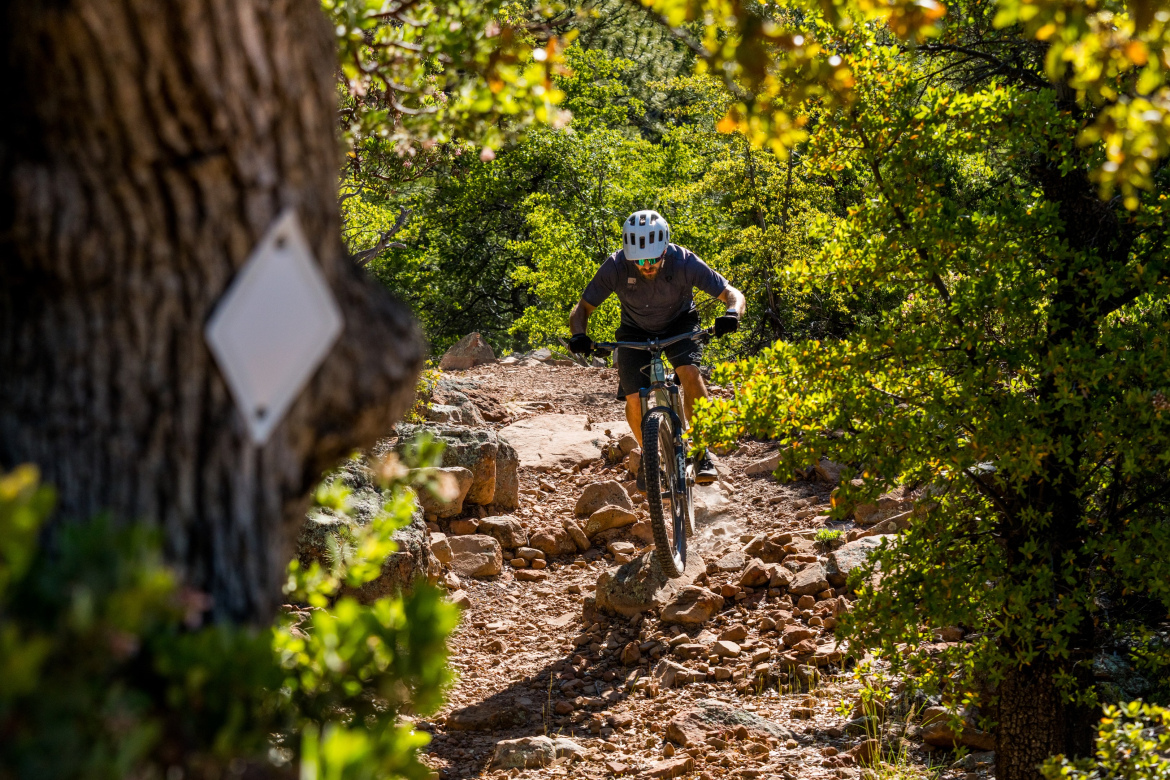 A mountain biker navigating a rocky trail through a forested area, with trees and foliage surrounding the path. The biker is wearing a helmet and is mid-ride, with the front wheel lifted slightly off the ground. Highline Trail: Geronimo's Call mountain bike trail.