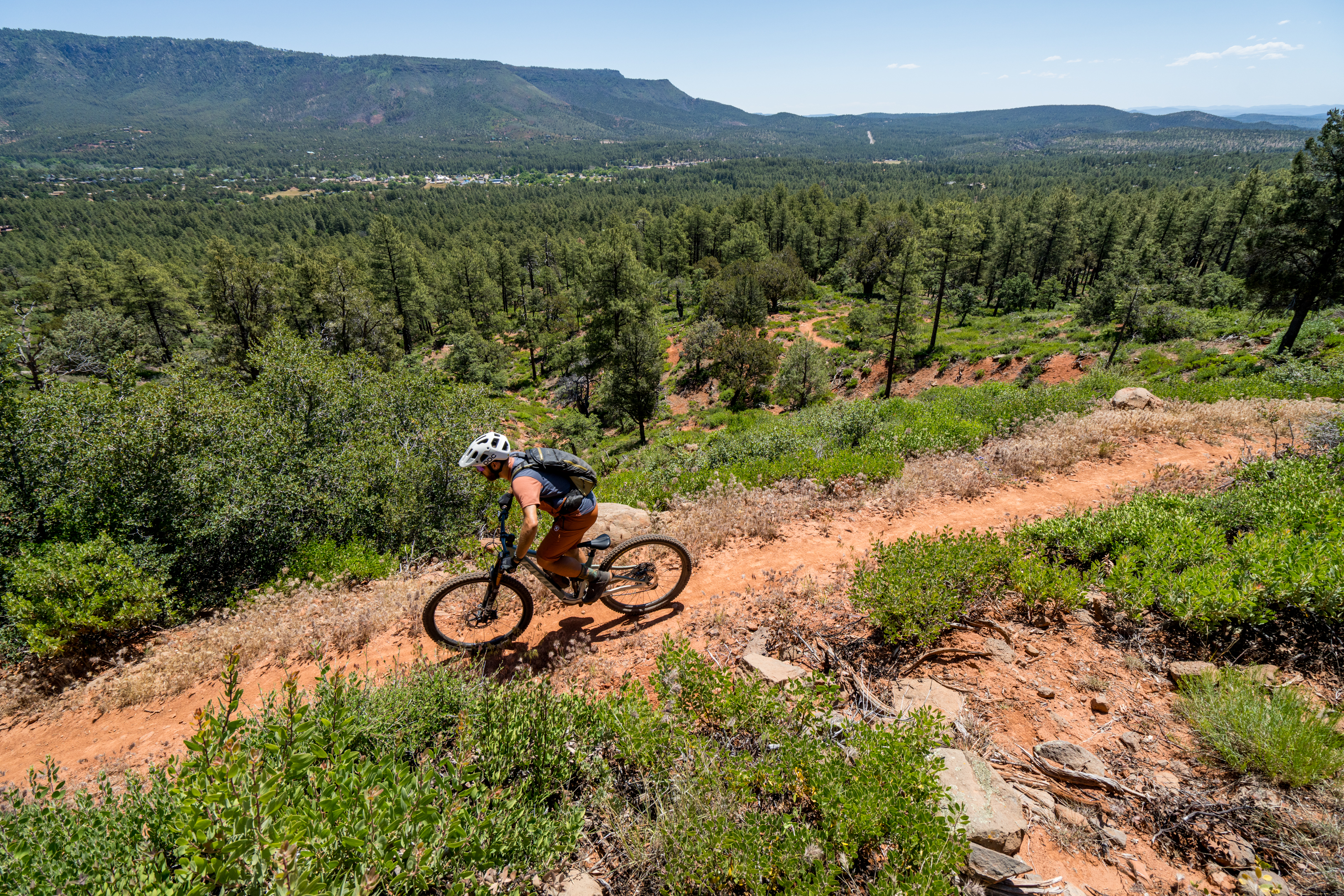 A mountain biker navigates a winding dirt trail on a sunny day, surrounded by lush greenery and distant mountains. The scene showcases a clear blue sky and clear visibility of the landscape, emphasizing the adventurous spirit of outdoor cycling.