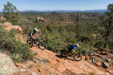 Two mountain bikers navigating a rocky trail through a forested landscape. The scene features rolling hills and boulders in the background, with clear blue skies overhead. One biker is in the foreground, focusing on the terrain, while the other rides slightly downhill behind him. The area is lush with greenery and surrounded by a mix of pine trees and rocky outcrops, showcasing an adventurous outdoor setting. Granite Dells mountain bike trail.