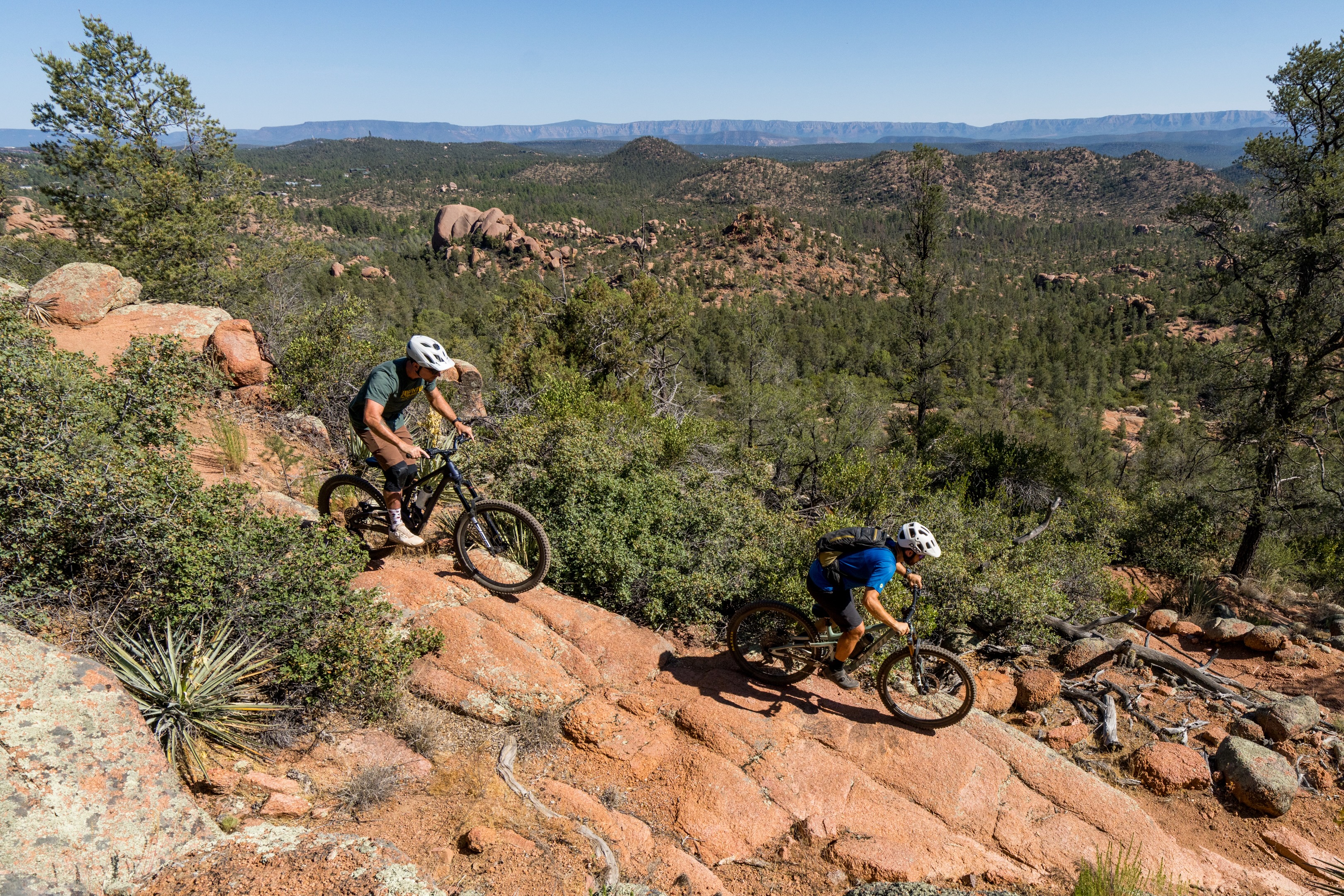 Two mountain bikers navigating a rocky trail through a forested landscape. The scene features rolling hills and boulders in the background, with clear blue skies overhead. One biker is in the foreground, focusing on the terrain, while the other rides slightly downhill behind him. The area is lush with greenery and surrounded by a mix of pine trees and rocky outcrops, showcasing an adventurous outdoor setting. Granite Dells mountain bike trail.