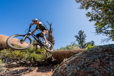 A mountain biker performing a jump over a rocky terrain with blue skies in the background. The cyclist is wearing a helmet and protective gear, showcasing an action-packed outdoor moment amidst a natural landscape. Granite Dells mountain bike trail.