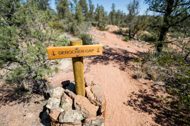 Wooden signpost marking the way to "Derocher Gap" with an arrow pointing downwards, located on a dirt trail surrounded by trees and shrubs in a natural outdoor setting. Granite Dells mountain bike trail.
