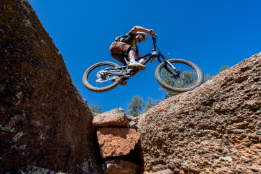 A mountain biker performing a jump over a gap between large rocks under a clear blue sky. Granite Dells mountain bike trail.