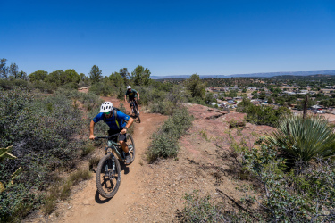 Two mountain bikers navigate a winding dirt trail through a hilly landscape, surrounded by vegetation, with a view of a small town below. The sky is clear and blue, indicating a sunny day. Ubuntu Trail mountain bike trail.