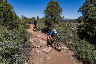 Two mountain bikers navigate a rocky trail surrounded by greenery under a clear blue sky. The riders are focused on their path, with one in a blue shirt and the other in a green shirt, as they make their way down a winding trail with scattered rocks. In the background, a scenic view of a nearby town and mountains can be seen. Ubuntu Trail mountain bike trail.