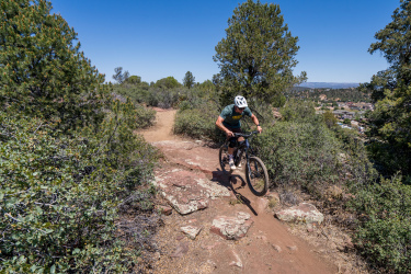 A person riding a mountain bike on a rocky trail surrounded by greenery and trees under a clear blue sky. The cyclist is navigating the terrain, showcasing an active outdoor lifestyle. In the background, a view of a town can be seen nestled among the hills. Ubuntu Trail mountain bike trail.