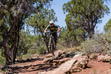 A mountain biker riding over rocky terrain on a trail surrounded by trees. The cyclist is wearing a helmet and protective gear, focusing on navigating the uneven ground. Dust is kicked up behind the bike as it moves swiftly through the natural landscape. Ubuntu Trail mountain bike trail.