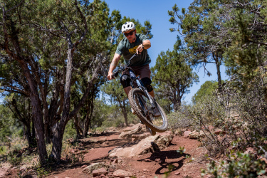 A mountain biker in a green shirt and helmet performs a jump on a rocky trail surrounded by trees under a clear blue sky. Ubuntu Trail mountain bike trail.