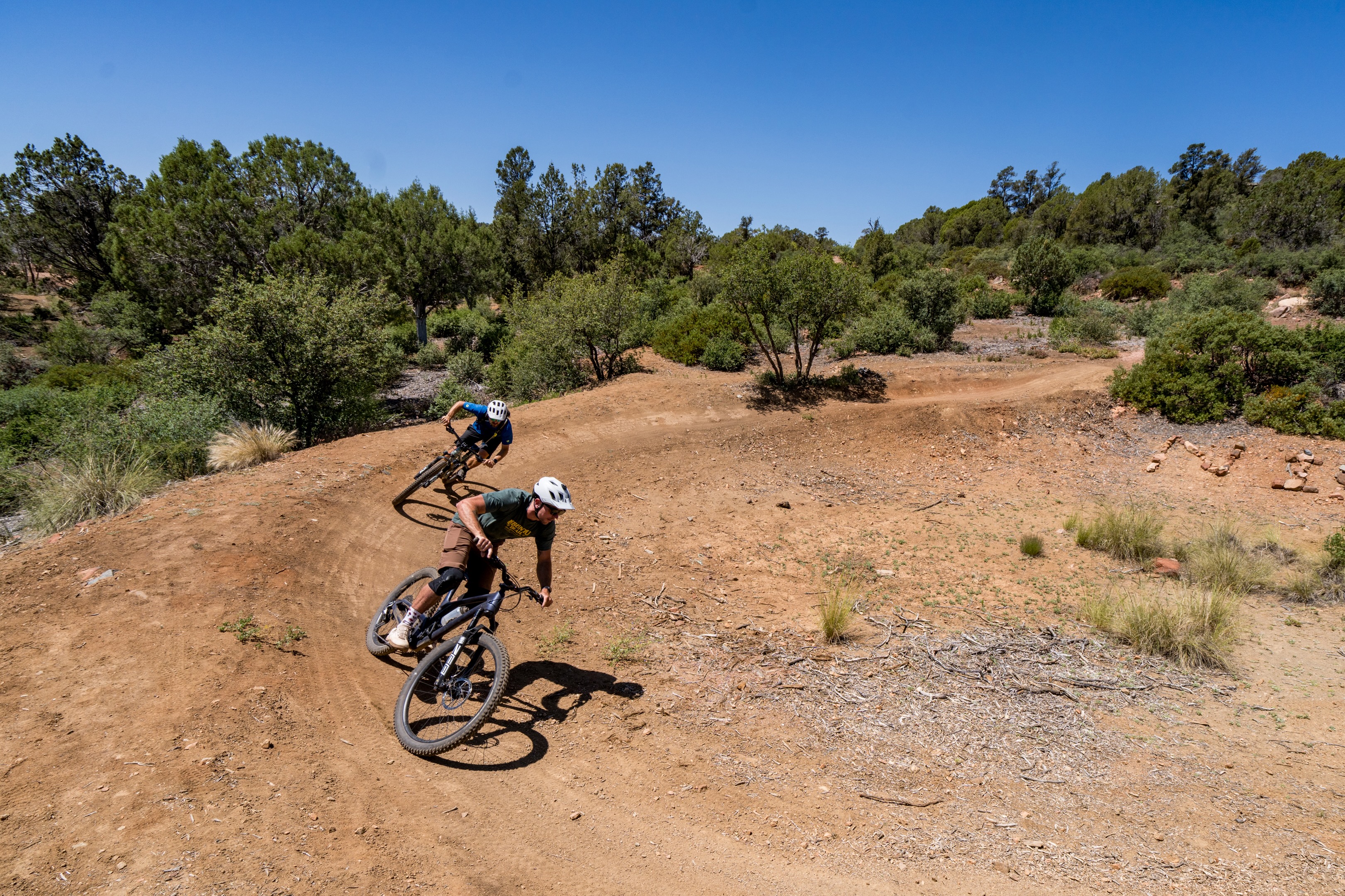 Two mountain bikers navigate a curved dirt trail surrounded by trees and shrubs under a clear blue sky. The riders are leaning into the turns, showcasing their skills and the movement of the bikes on the rugged terrain. Ubuntu Trail mountain bike trail.
