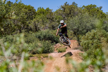 A mountain biker performing a jump on a dirt trail surrounded by greenery. The biker is mid-air, showcasing an action pose. The bright blue sky is visible above, emphasizing a sunny day. Ubuntu Trail mountain bike trail.
