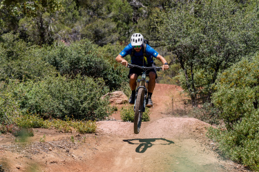 A mountain biker in a blue shirt and helmet performs a jump on a dirt trail surrounded by greenery. Ubuntu Trail mountain bike trail.