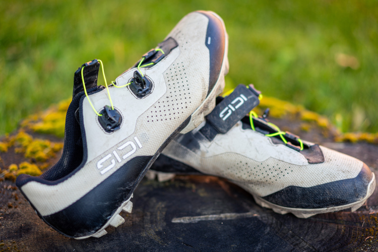 A pair of SIDI cycling shoes resting on a wooden surface, featuring a predominantly white and black design with neon green laces. The shoes appear slightly worn, suggesting they have been used for cycling activities. The background shows a blurred grassy area, adding a natural outdoor context.