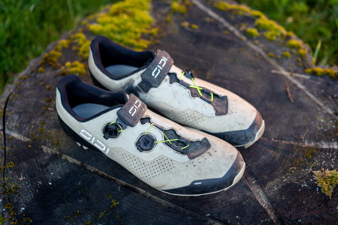 A pair of worn cycling shoes with a sleek design, featuring a predominantly white exterior and black accents. The shoes have a dirtied appearance, highlighting their use, and are secured with a closure system that includes visible laces and clips. They are positioned on a moss-covered wooden surface, with greenery in the background.