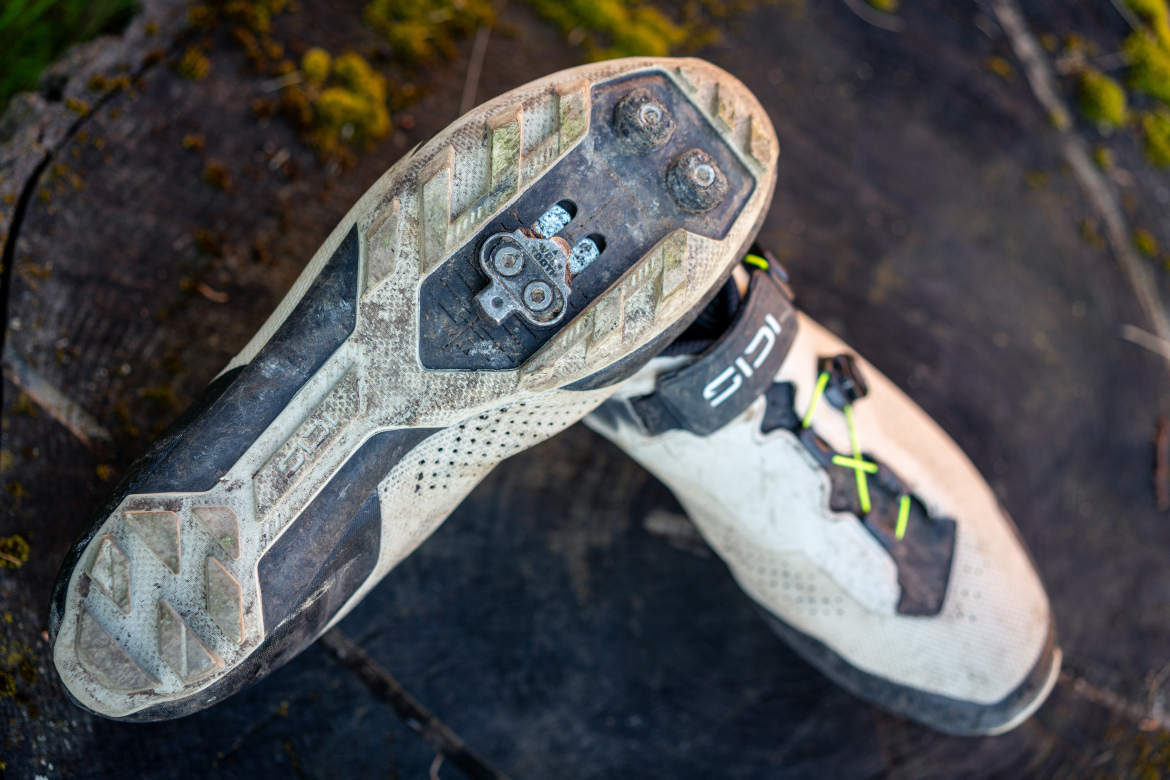 Alt text: A pair of worn cycling shoes with cleats on a wooden surface, showcasing a predominantly white design with black and neon yellow accents. The soles are dirty, indicating recent use.