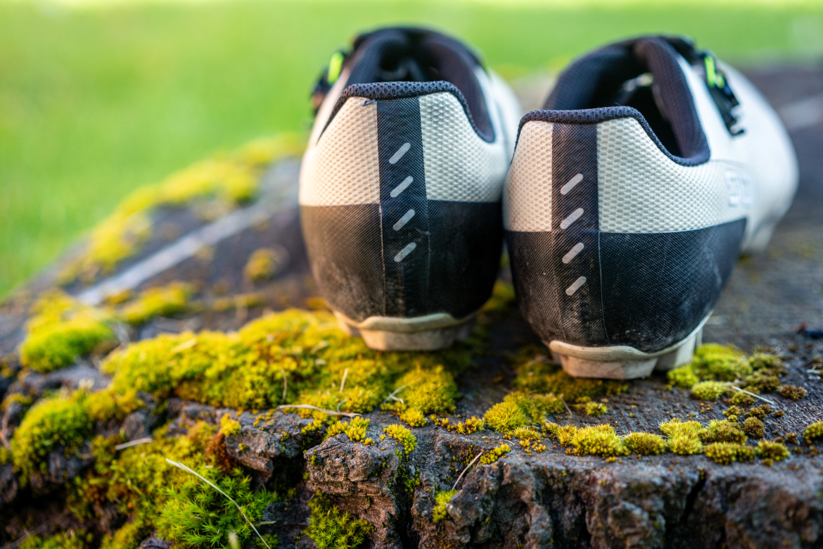 A close-up view of the back of two athletic shoes resting on a moss-covered wooden surface, with a blurred green background. The shoes feature a mix of black and white materials and have detailing on the heel.