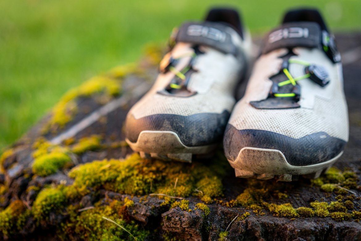 Close-up of a pair of hiking shoes resting on a moss-covered log. The shoes feature a white and gray design with black accents and bright green laces. The outdoor setting suggests a connection to nature and adventure.